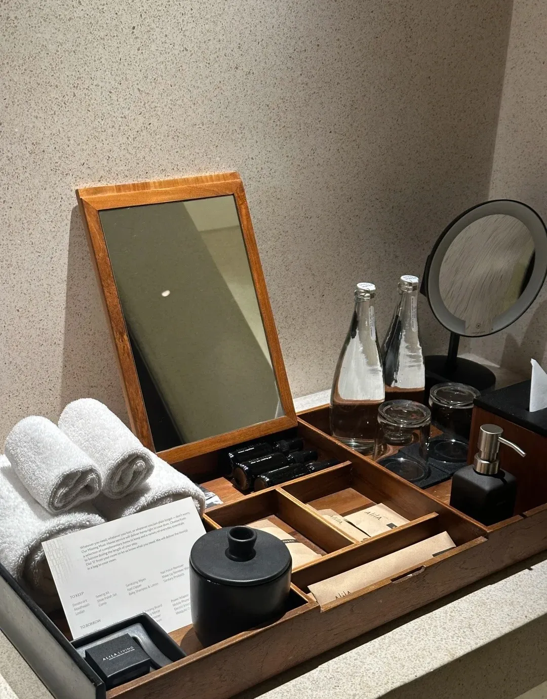 A wooden vanity tray with rolled towels, toiletries and glass water bottles