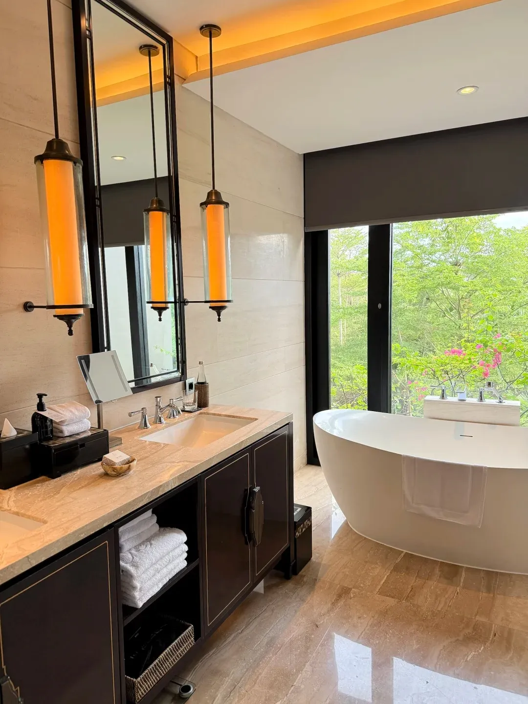 A Kempinski bathroom with a double vanity, hanging lanterns and a freestanding tub by the window
