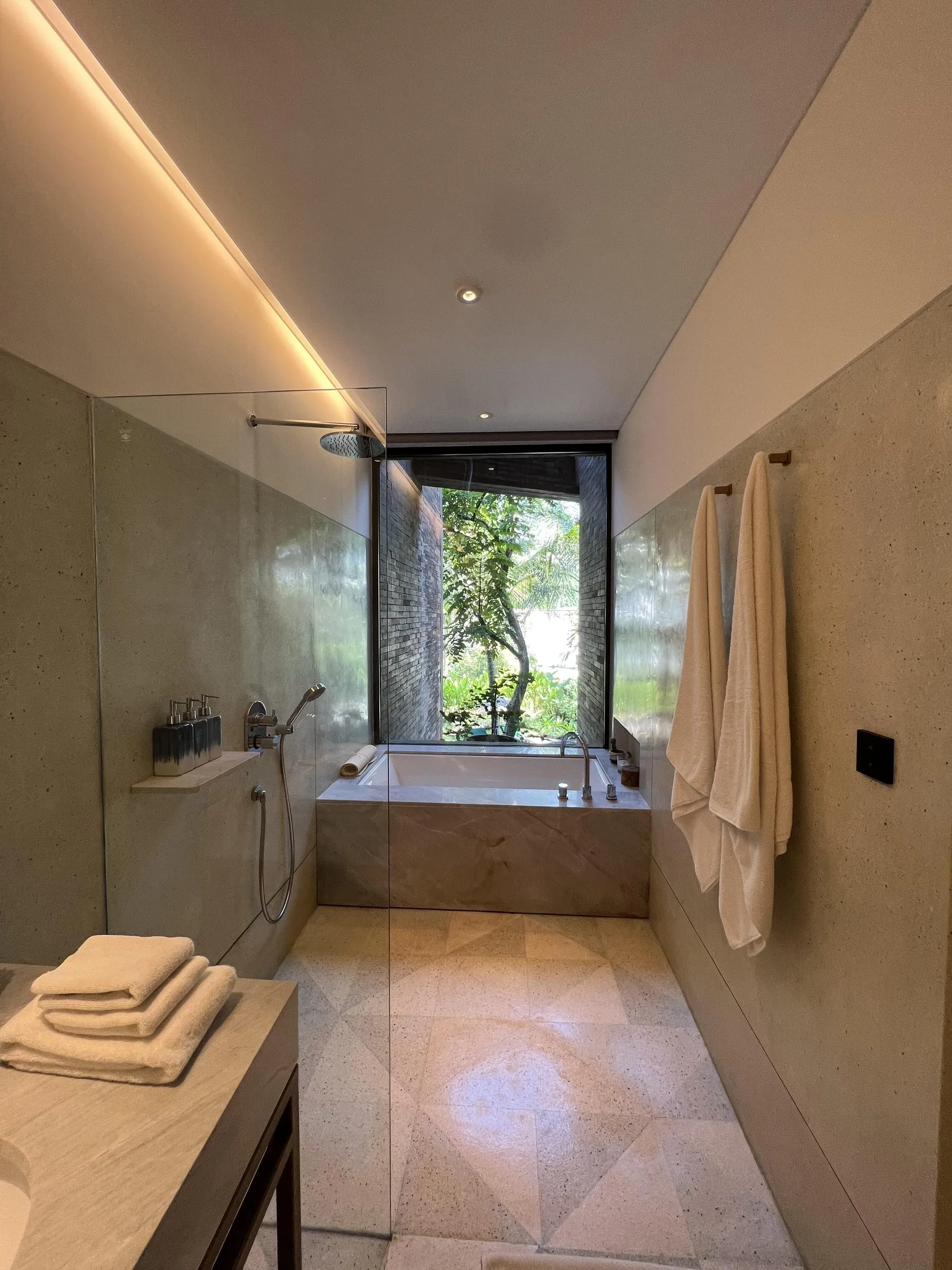 A long stone bathroom with a walk-in rain shower and a deep tub set under a tall window onto the garden