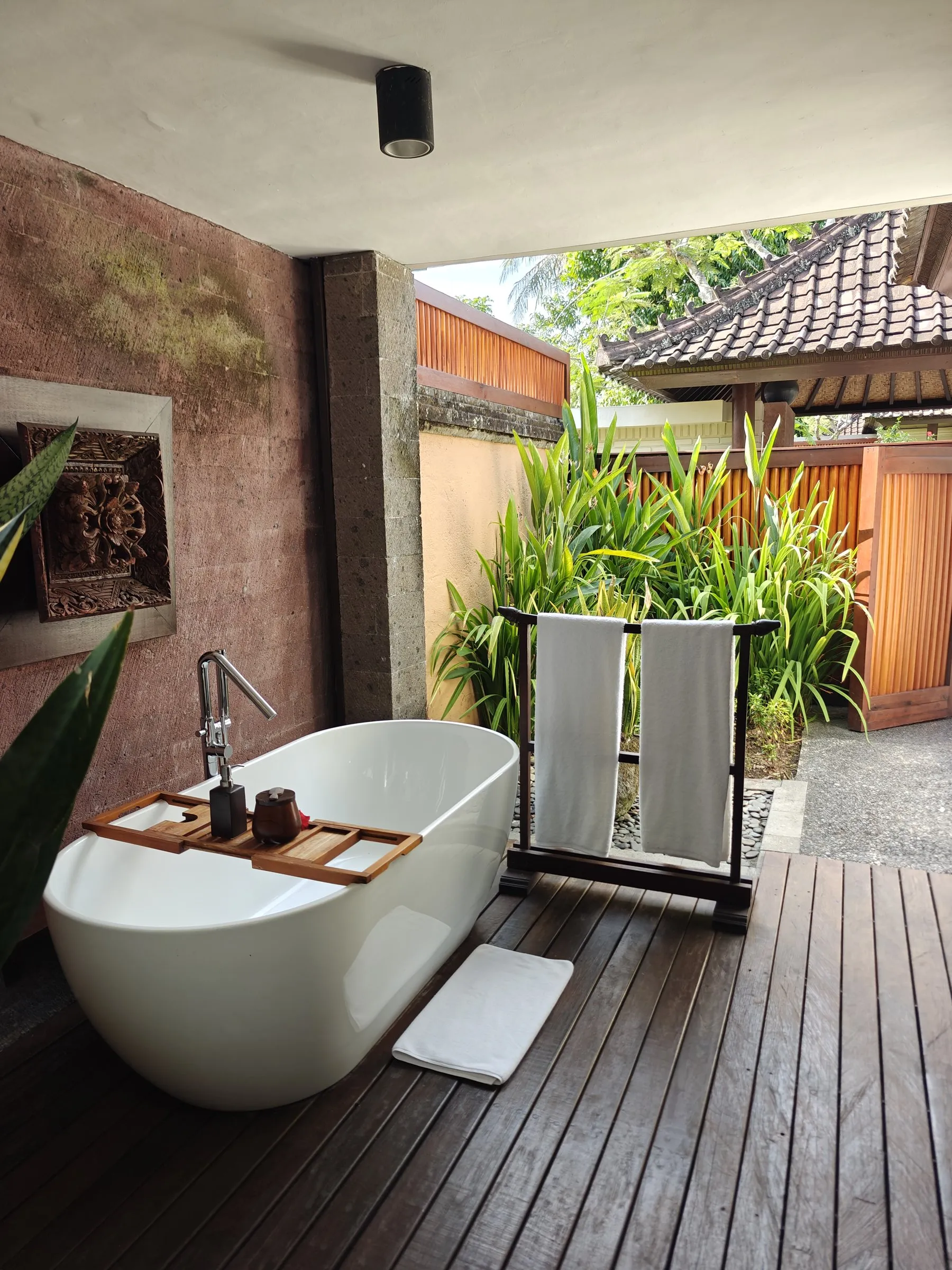 A freestanding bathtub on an open-air timber deck, with a garden and a thatched roof beyond