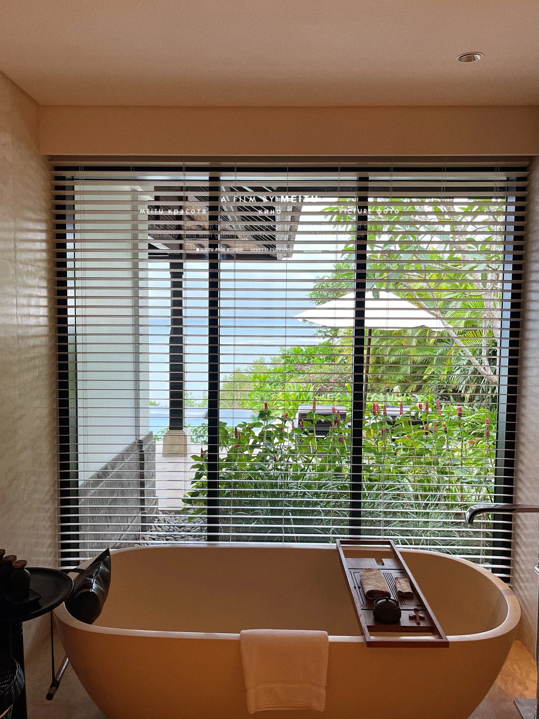 A freestanding bathtub in front of a tall window with half-open blinds and tropical garden outside