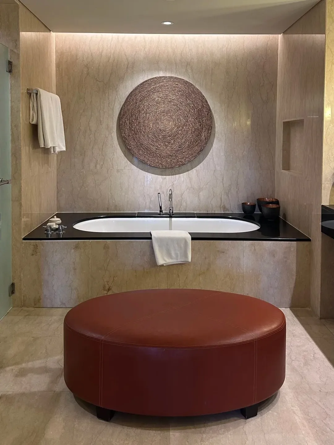A built-in bathtub in a marble bathroom, with a leather ottoman in front