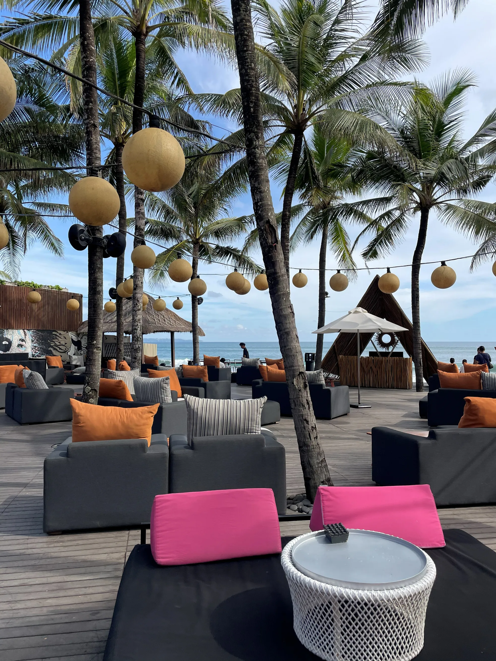 Lounge sofas with orange cushions under palms and strung lanterns on a deck by the sea, with a triangular W booth on the beach