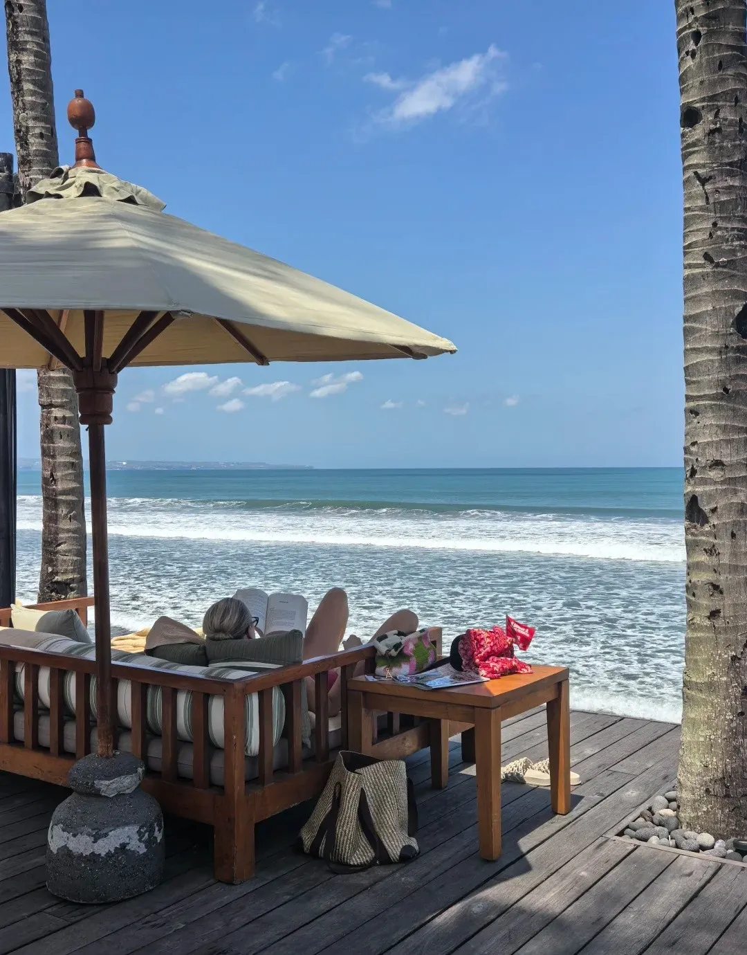 A daybed under an umbrella on a timber deck right above the beach and the waves