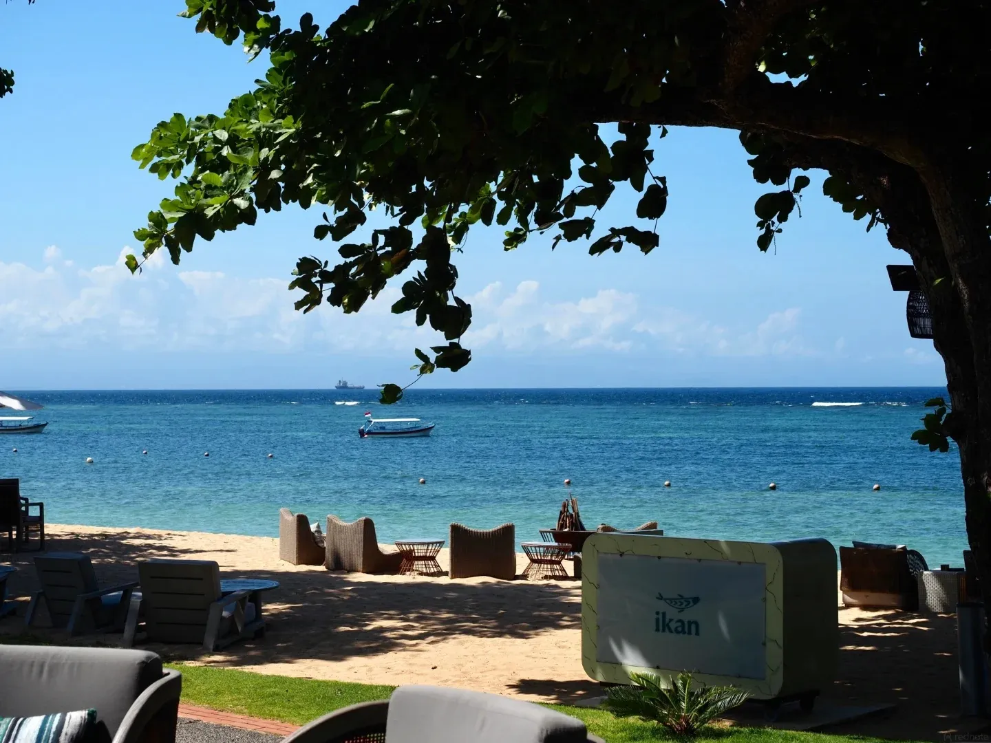 Calm blue water and boats seen from under a shady tree on the beach