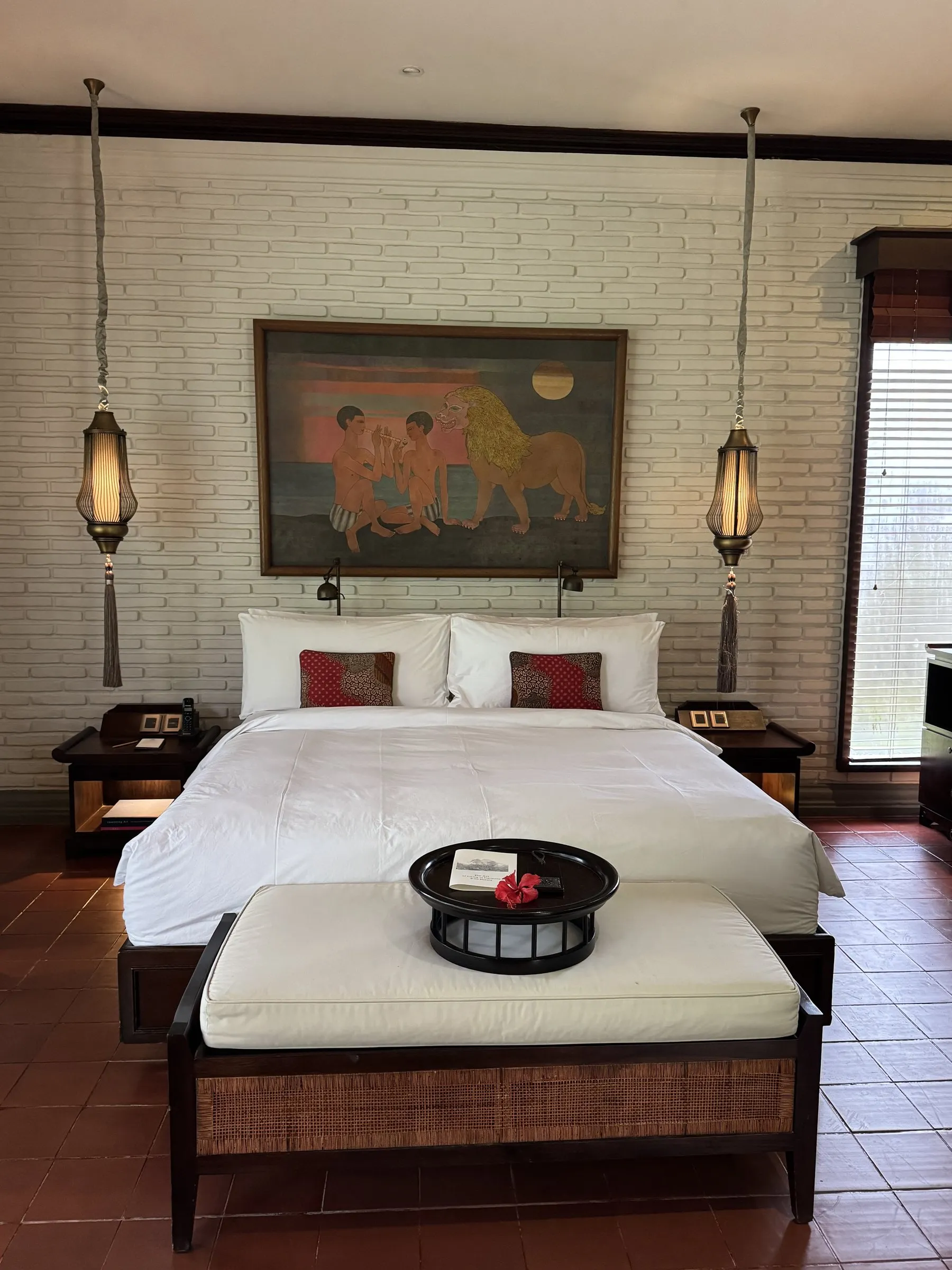A bedroom with a Balinese painting over the bed, hanging brass lanterns, white brick walls and terracotta floors