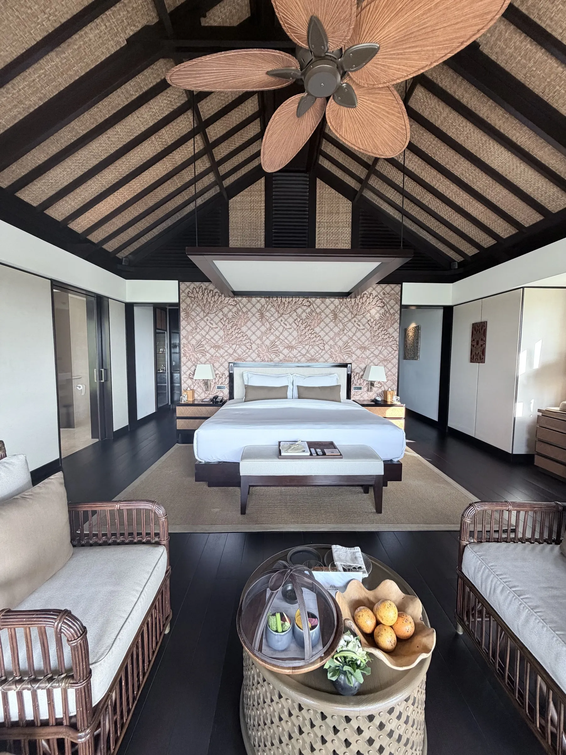 A Raffles Bali villa bedroom under a high thatched roof with a palm-leaf fan, a king bed and rattan daybeds around a fruit table