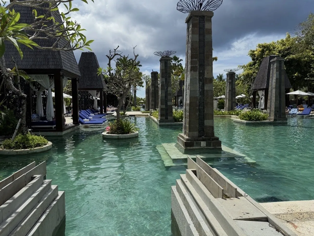 The pool’s stone columns and curtained cabanas, with blue loungers along the water