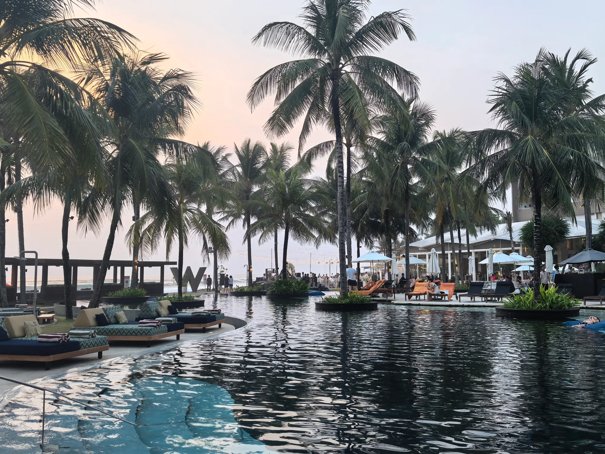 The pool at dusk, with daybeds on the edge, palms on planted islands and the W sign on the beach beyond