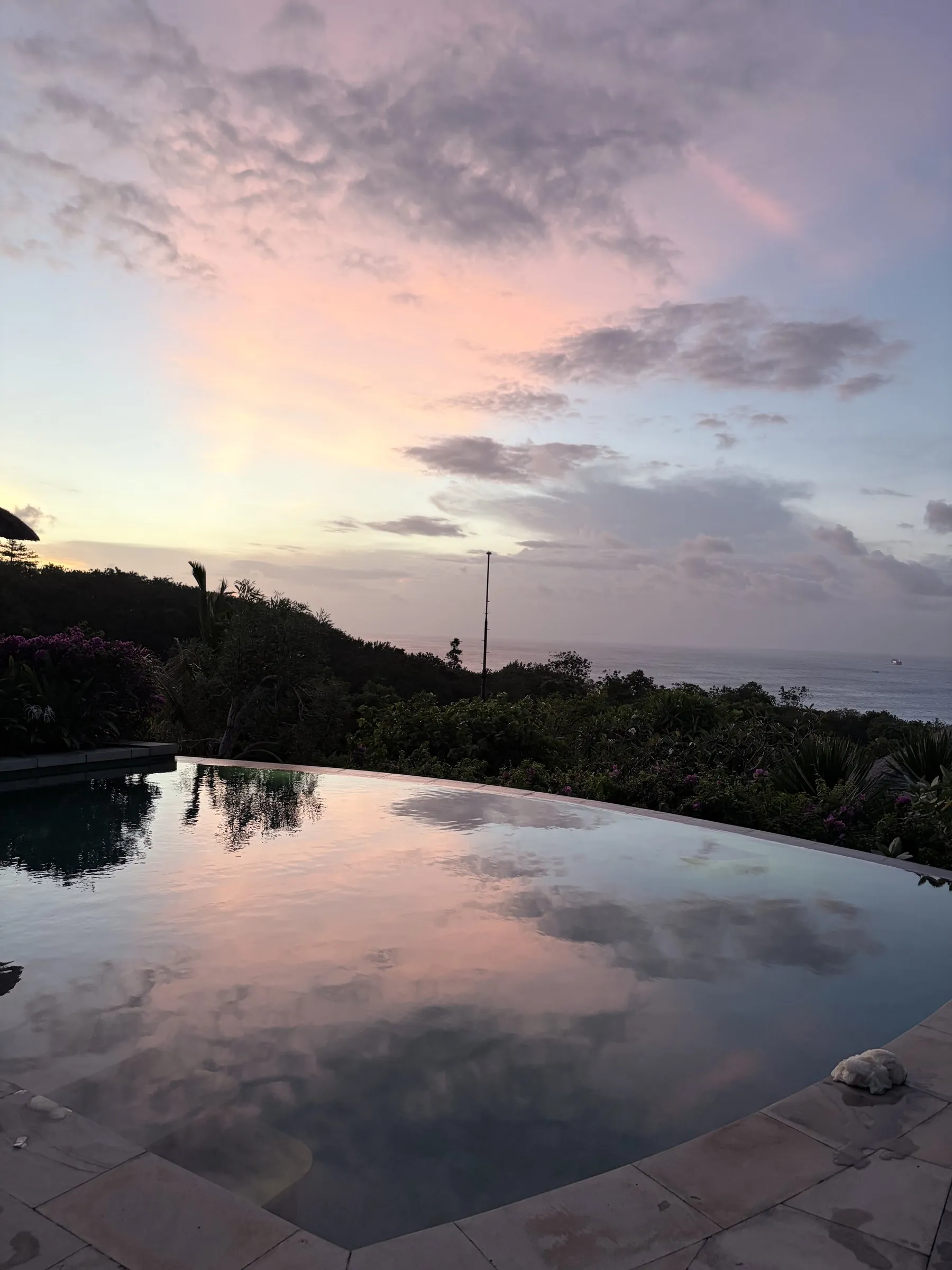 The infinity pool reflecting a pink sunset sky, with the hillside and the sea behind