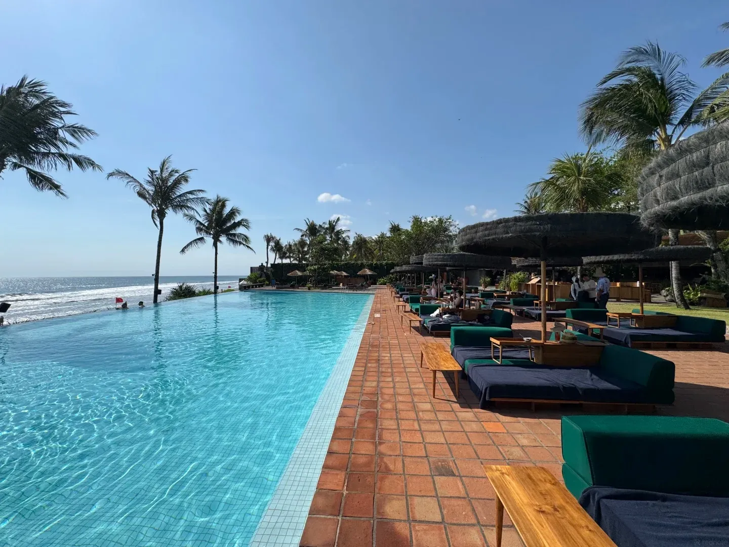 The infinity pool running along the beachfront, with green daybeds and thatched umbrellas on the tiled deck