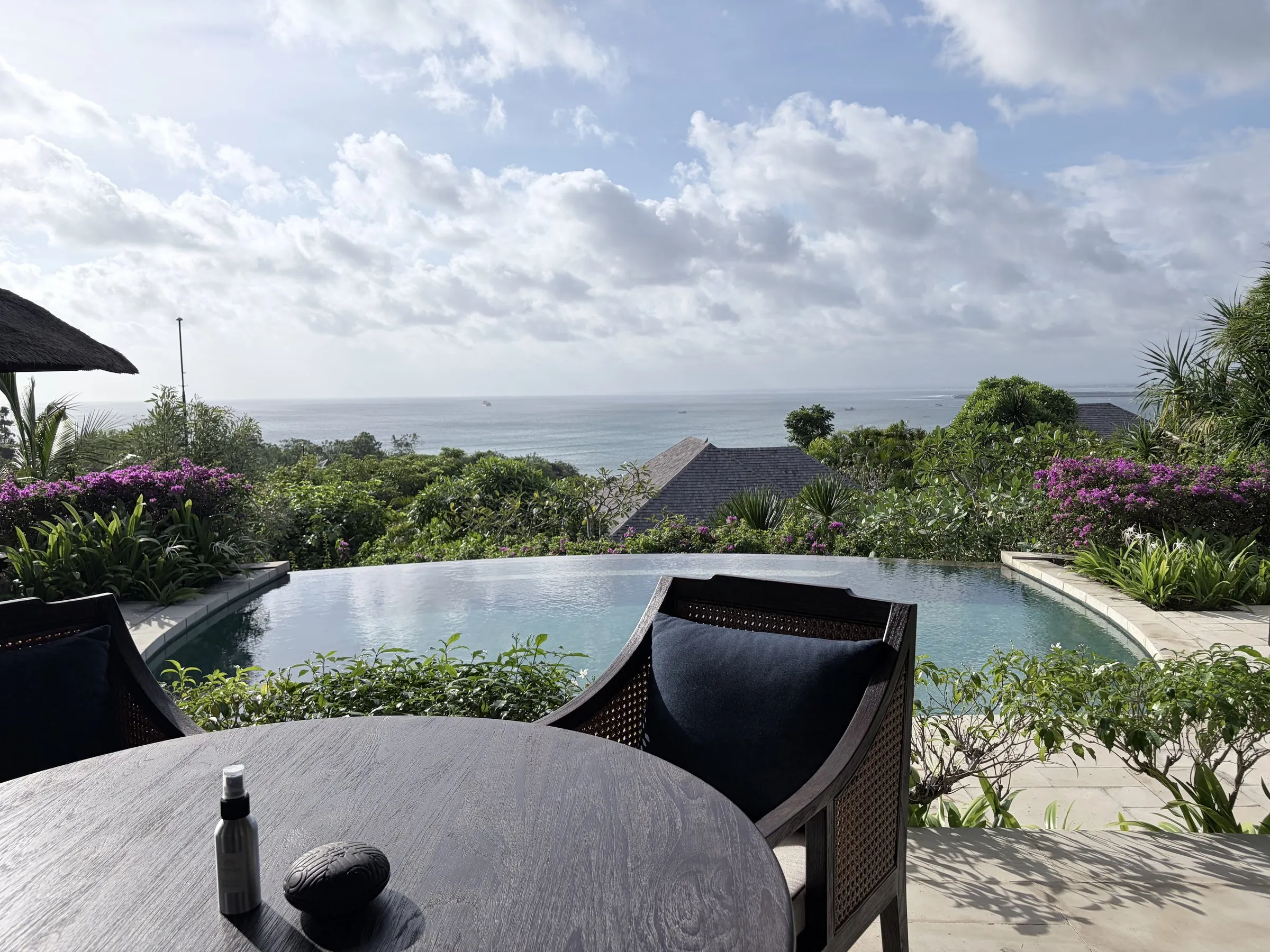 A table and chairs above a curved infinity pool, with gardens and villa roofs running down to Jimbaran Bay