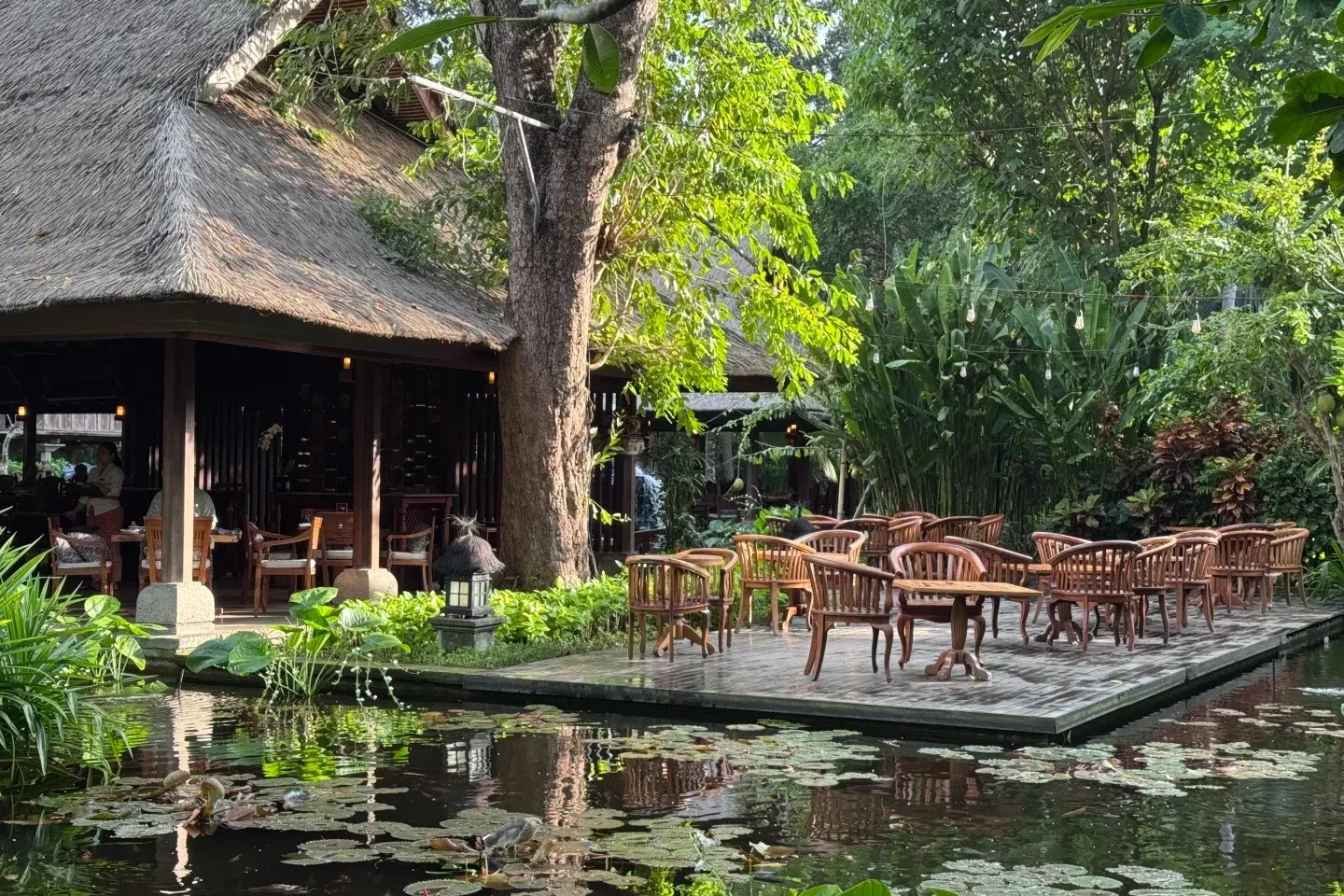 An open-air restaurant terrace under a thatched roof, set out over a lily pond