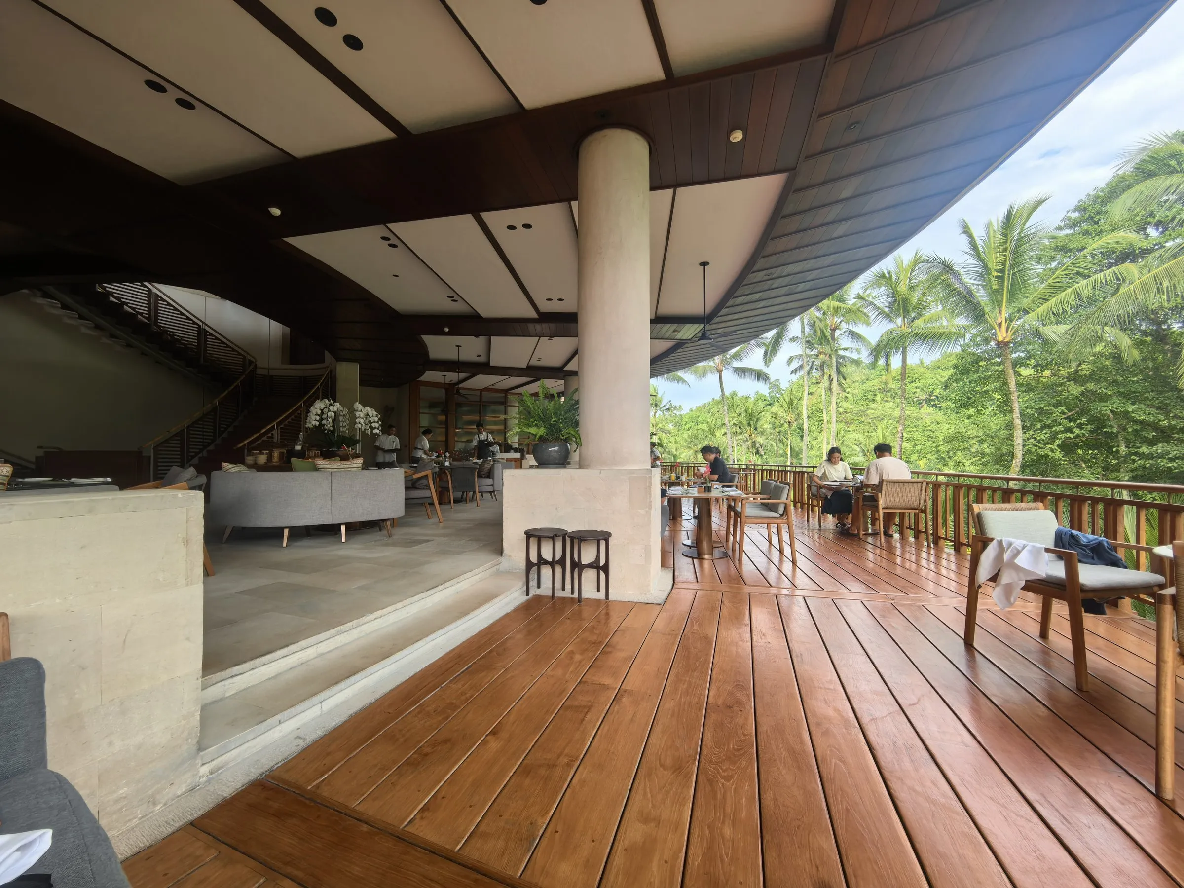 A restaurant terrace with a timber deck under a sweeping roof, open to the palms