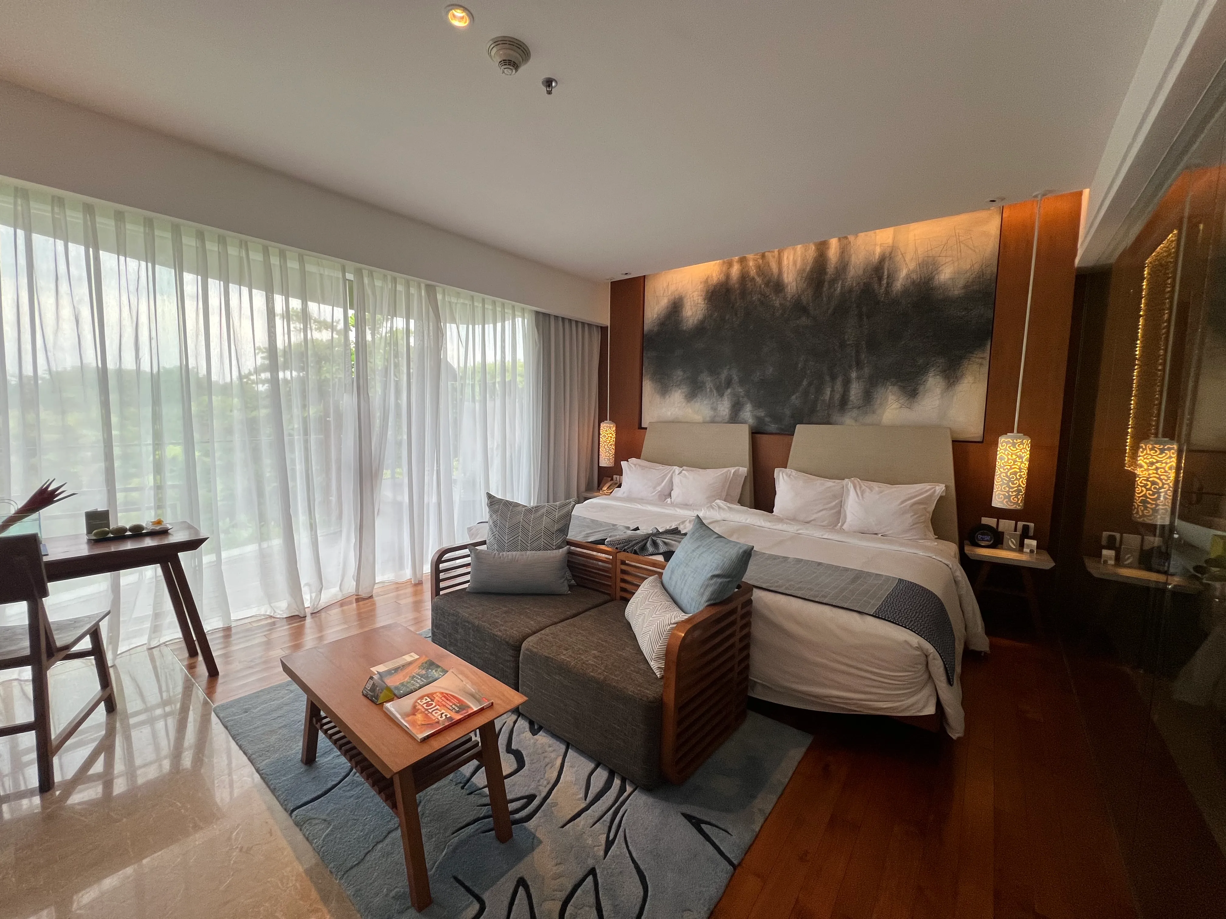 A Maya Sanur room with two beds under a large ink painting, a sofa, a desk and sheer curtains over a balcony onto the trees