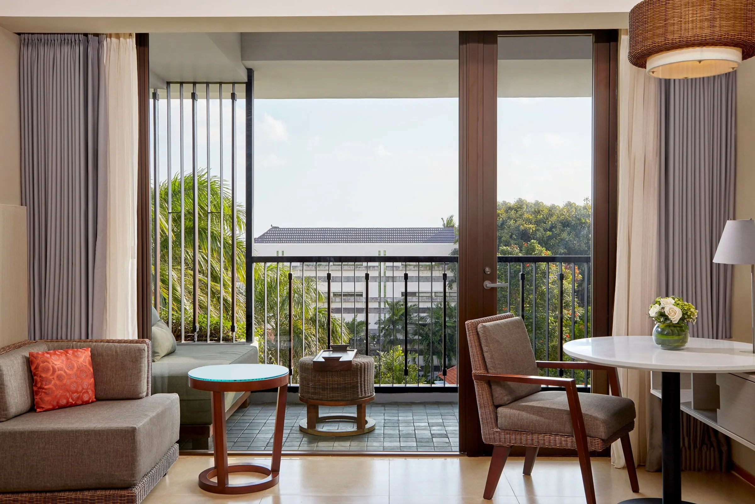 A Courtyard Seminyak room with a sofa, armchair and round table by glass doors onto a balcony with a daybed