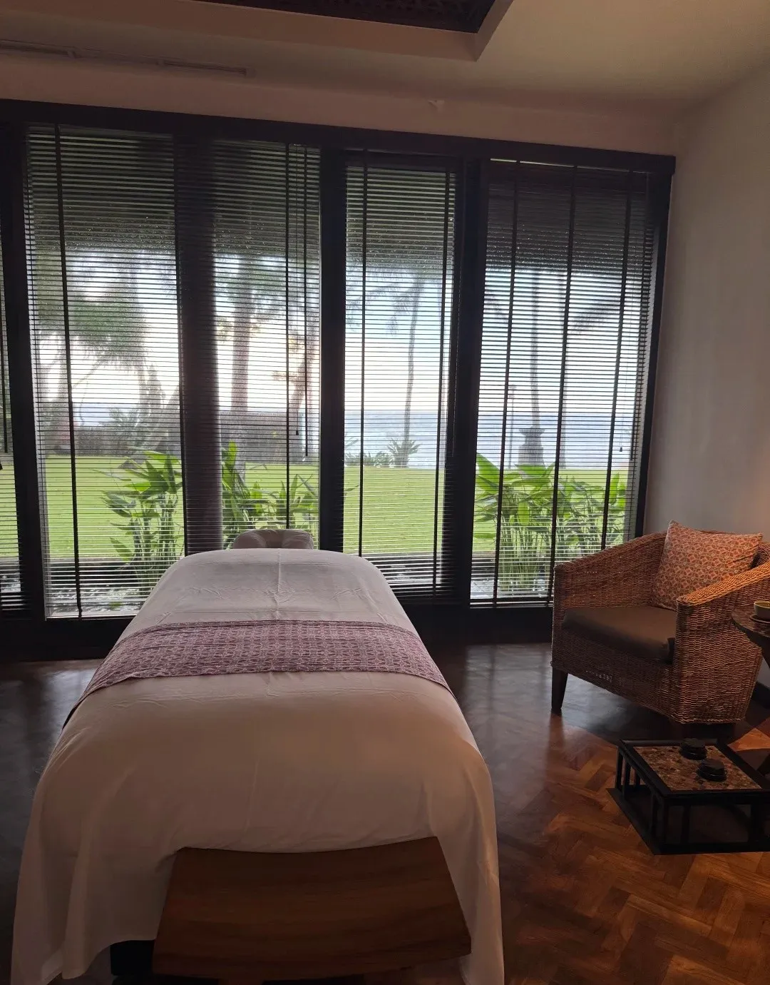 A spa treatment room with a massage bed and tall windows onto the lawn and the sea