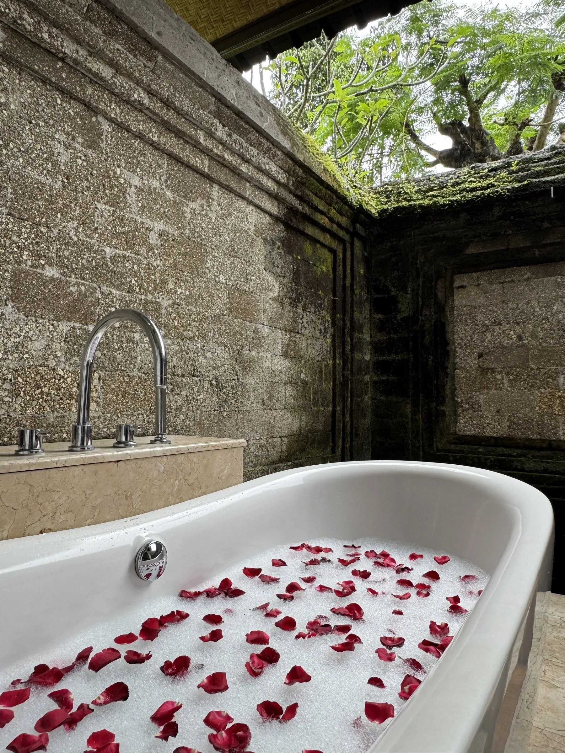 A bath scattered with rose petals inside mossy stone walls, open to the sky and the trees