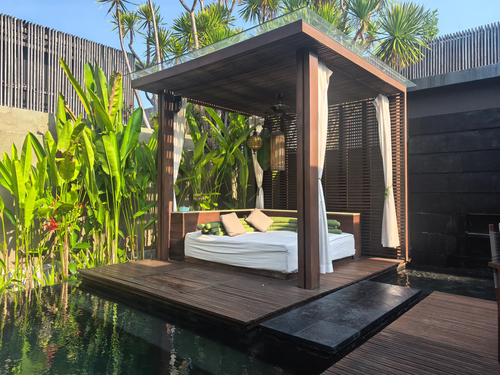 A villa’s private pool with a curtained daybed pavilion on a timber deck, walled in by tropical plants