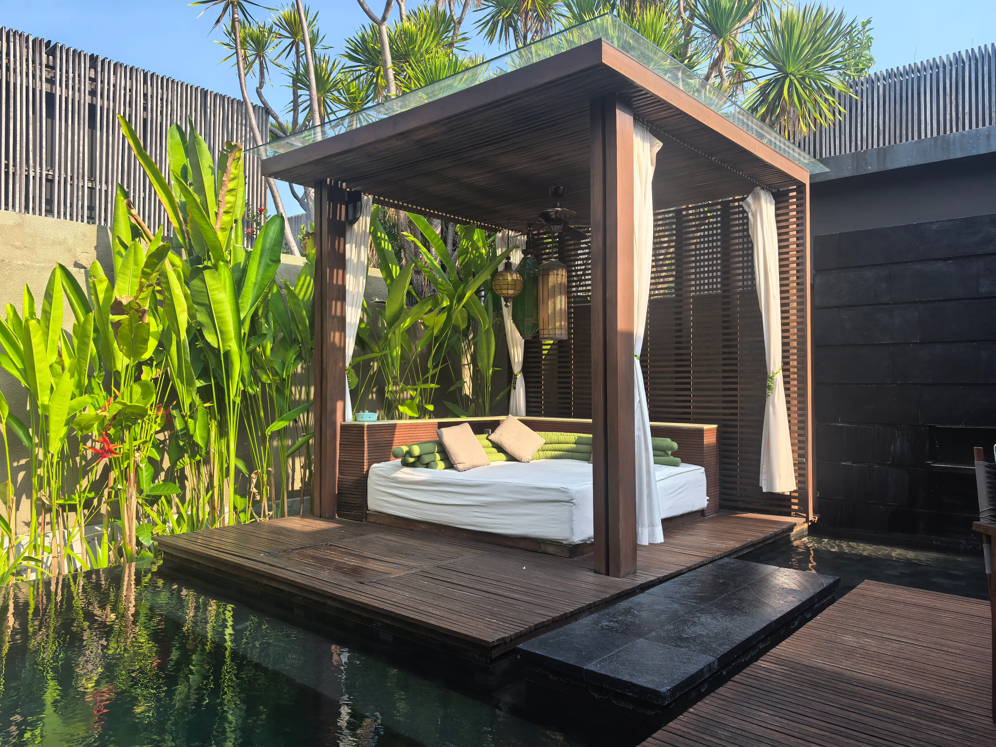 A villa’s private pool with a curtained daybed pavilion on a timber deck, walled in by tropical plants
