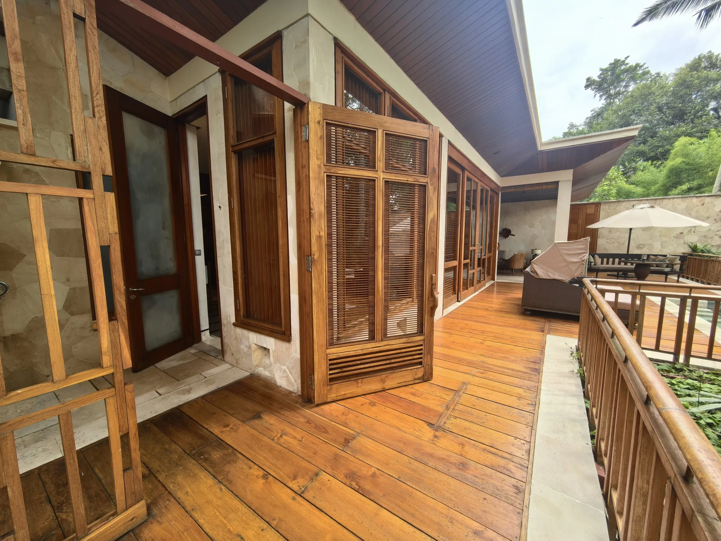 A timber deck running along a suite past carved shutters, with an umbrella and the jungle beyond