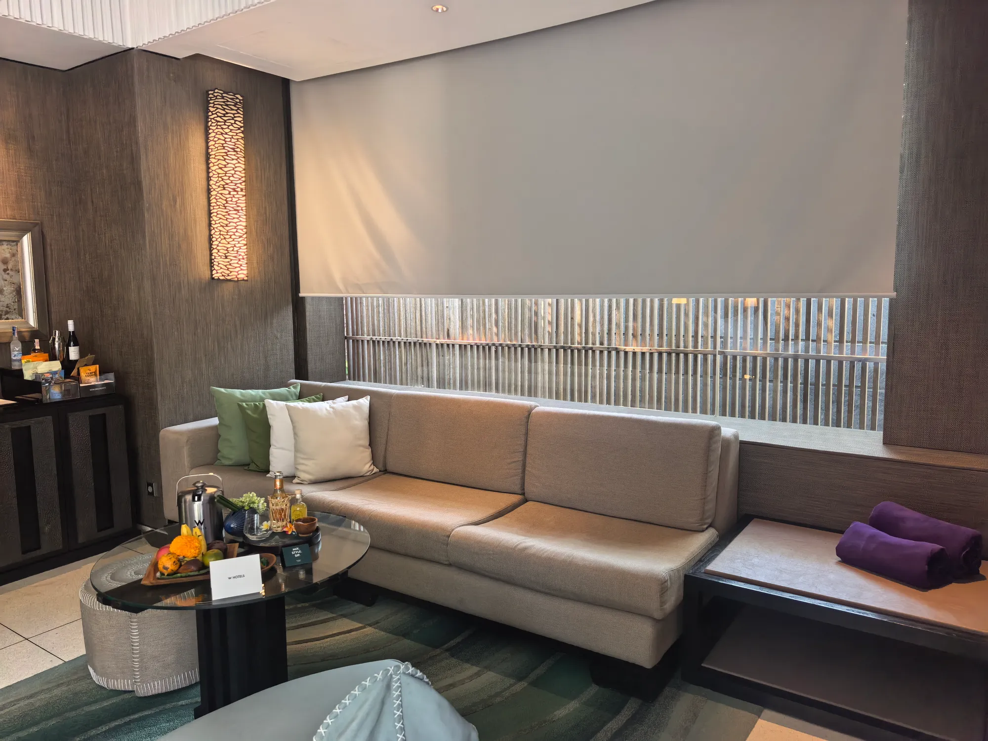 A villa living room with a long sofa under a slatted window, a fruit platter on the coffee table and a minibar