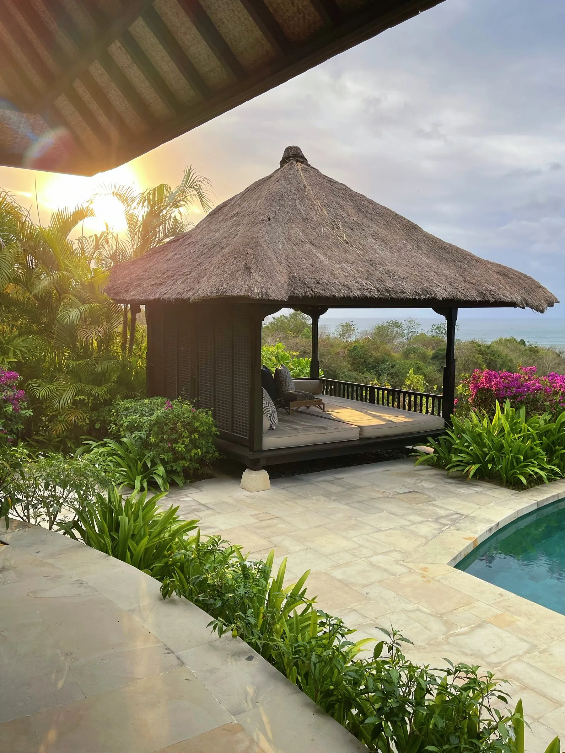 A thatched daybed pavilion beside a villa’s private pool, with bougainvillea and a glimpse of the sea behind